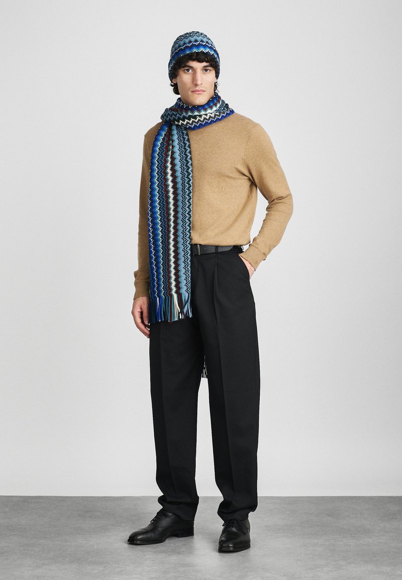 Knit hat and scarf set in blue tones with zigzag patterns. Brown sweater and black trousers. Smooth texture and fringed scarf ends.