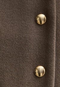 Bruin wolweefsel met een zachte textuur, voorzien van twee glanzende gouden knopen als accenten. Het weefsel heeft een subtiele, gelijkmatige korrelpatroon.