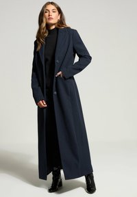 Donna che indossa un lungo cappotto blu scuro sopra un dolcevita nero e pantaloni, in piedi con una mano nella tasca del cappotto e stivali neri con tacco.