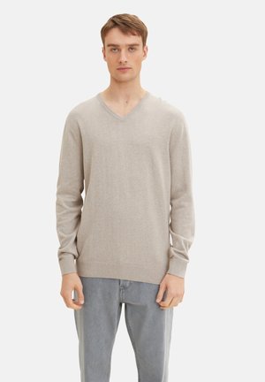 Beiger V-Ausschnitt-Pullover aus weichem, leichtem Stoff mit gerippten Bündchen und Saum. Kombiniert mit hellgrauen Jeans. Minimales Logo-Detail auf der Brust.