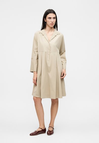 Γυναίκα στέκεται φορώντας ένα μπεζ φόρεμα μήκους έως το γόνατο με κουμπιά, μακριά μανίκια και σκούρα καφέ φλατ παπούτσια με λουράκια στον αστράγαλο.