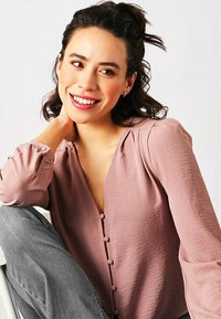 Camicetta rosa con bottoni, maniche lunghe, tessuto strutturato e scollatura a V. La camicetta presenta una vestibilità comoda e decorazioni con bottoni.