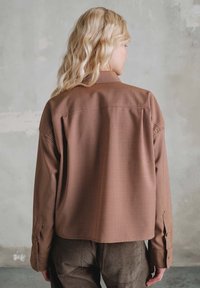 Blondynka z falującymi włosami stojąca tyłem, ubrana w brązową, w kratkę koszulę z długim rękawem i ciemne spodnie z sztruksu na tle szarej ściany.