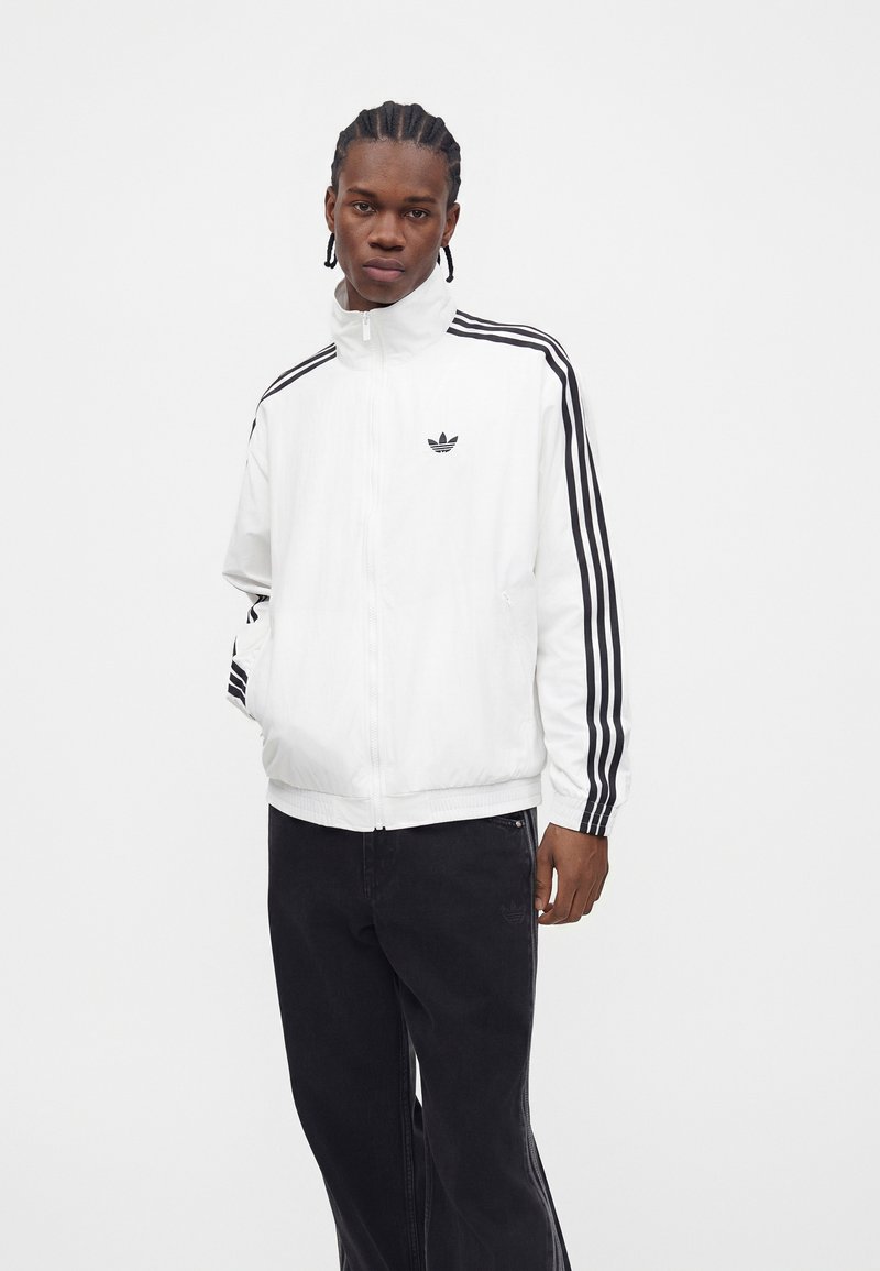 Ung mand med fletninger, iført en hvid Adidas-jakke med sorte striber og sorte bukser, stående med den ene hånd i lommen.