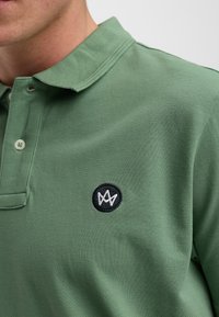 Grön polo skjorta i texturerad bomull, med krage, två knappar och en rund svart broderad logo på vänster bröst.