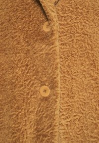 Bruine fleecestof met een gestructureerd oppervlak, voorzien van een enkele lichtbruine knoop en een reverskraagdesign.