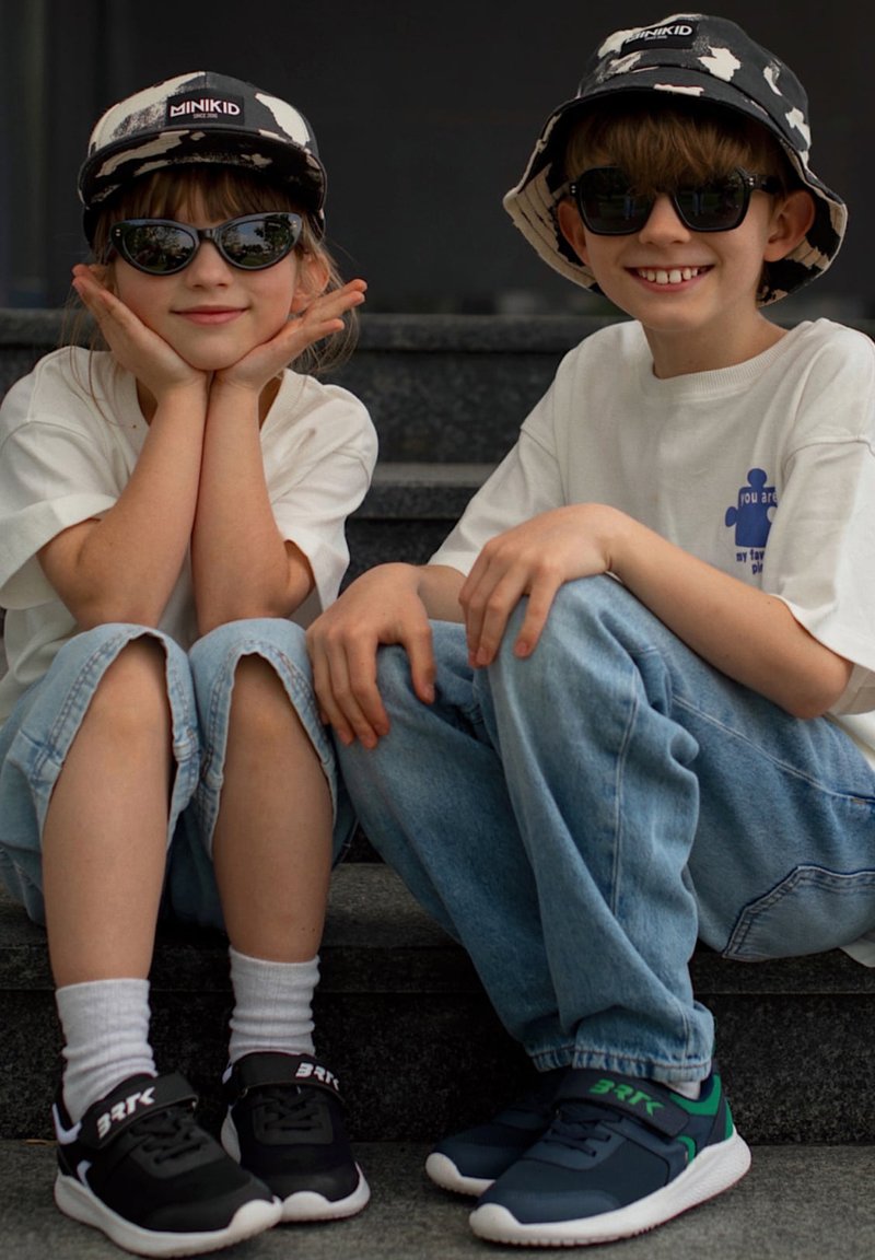 Deux enfants portant des chapeaux noir et blanc assortis et des lunettes de soleil sont assis sur des marches, souriants ; ils portent tous les deux des chemises blanches et des chaussures décontractées.
