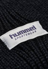 Svart stickad tyg med en vit etikett som har lila text som läser "Hummel SPORTSWEAR". Texturen verkar mjuk och strukturerad.