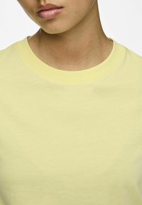 Tricou din bumbac de culoare galben deschis, cu decolteu rotund, textură netedă și detalii minime de cusătură pe guler.