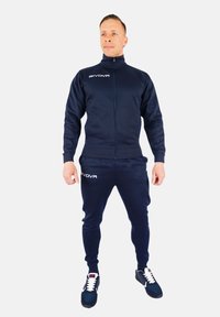 Mornarska zip-up trenirka z ujemajočimi se hlačami, izdelana iz mehkega materiala. Vključuje logo na prsih in stegnu; teksturirani manšeti in pas.