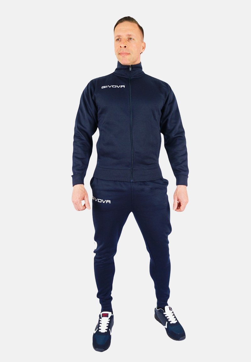 Mornarska zip-up trenirka z ujemajočimi se hlačami, izdelana iz mehkega materiala. Vključuje logo na prsih in stegnu; teksturirani manšeti in pas.