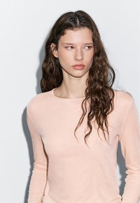 Helles rosa Langarmoberteil aus einem weichen, glatten Stoff. Rundhalsausschnitt mit schmaler Passform. Haare in lockeren, natürlichen Wellen frisiert.