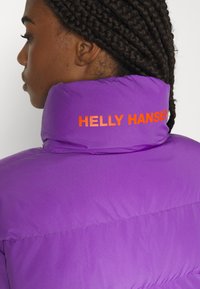 Violetinė pūkinė striukė su aukštu apykakle. Atgal yra drąsus oranžinis "HELly Hansen" logotipas. Lygus, blizgus audinys su pasiūtu raštu.