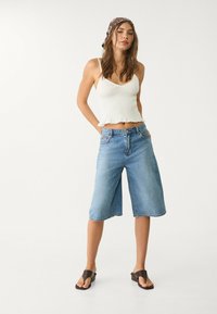 Culotte in denim blu chiaro con una vestibilità rilassata, orlo grezzo e tasche frontali. Abbinato a un top corto bianco in tessuto testurizzato e sandali marroni.