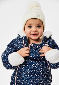 Marineblauwe donsjas met wit bloemmotief en pluche manchetten, gecombineerd met een crèmekleurige geribbelde gebreide muts met een pompon bovenop.