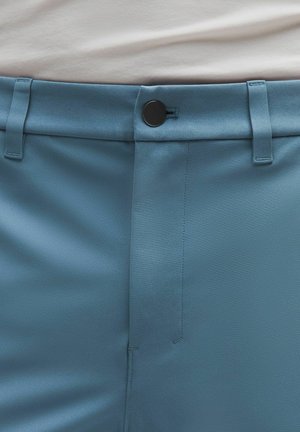 Detalle de la cintura de unos pantalones azules con un botón negro, trabillas para el cinturón y una camisa blanca parcialmente visible metida dentro.