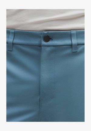 Detail der blauen Hose an der Taille mit einem schwarzen Knopf, Gürtelschlaufen und einem teilweise sichtbaren, hineingesteckten weißen Hemd.