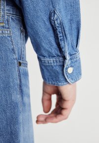 Jasnoniebieska koszula dżinsowa z widocznymi szwami, zapięciem na guziki i tkaninową metką blisko mankietu. Charakteryzuje się gładką teksturą.