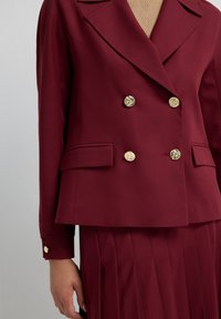 Burgundy double-breasted blazer with gold button accents, side pockets, and a structured fit, paired with a pleated skirt.