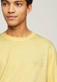 Camiseta de algodón amarilla con mangas cortas y cuello redondo. Sutil logotipo bordado de "Tommy" en el pecho. Textura suave y ajuste casual.