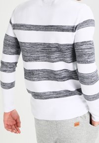 Haut blanc à manches longues avec de larges rayures horizontales gris charbon, porté par une personne portant un pantalon gris clair avec une petite étiquette brune indiquant "REBEL".