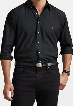 Uomo che indossa una camicia nera con bottoni e logo rosso, pantaloni neri, cintura nera con fibbia in argento e orologio da polso, braccia rilassate ai lati.