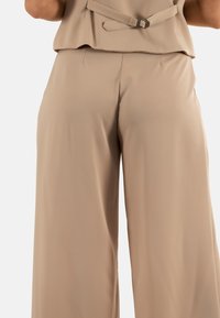Pantalons à jambe large en tissu beige doux, présentant une texture lisse et un détail de taille ceinturée, offrant une coupe décontractée.