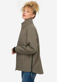Giacca softshell verde oliva con colletto alto, chiusura frontale con zip e polsini neri a contrasto. Presenta taschelaterali con zip e dettagli arancioni.