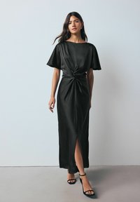 Fekete selyem maxi ruha rövid ujjakkal, csavart részlettel a deréknál és combig érő hasítékkal. Fekete pántos magassarkúval párosítva.