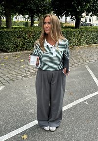 Striped green and white polo with a button collar, paired with high-waisted gray wide-leg trousers and gray slip-on shoes; holding a coffee cup.