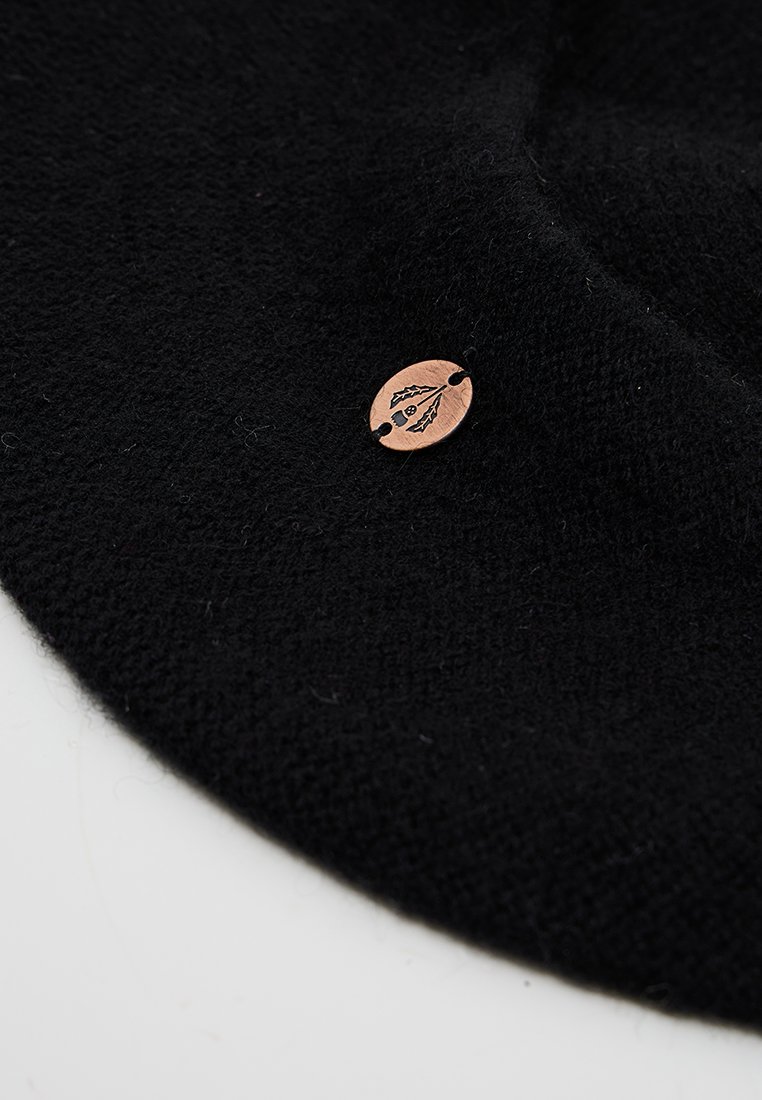 Czarna wełniana czapka o miękkiej teksturze, z małą, okrągłą miedzianą metką z wytłoczonym wzorem. Czapka prezentuje prosty, klasyczny kształt.