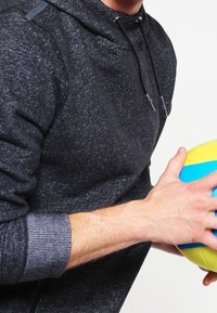 Felpa grigia con cappuccio, texture e dettagli neri, con polsini a costine; una persona tiene un pallone multicolore in una mano.