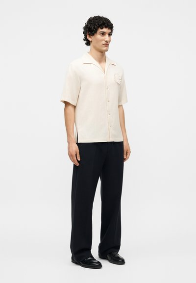Ένας άνδρας στέκεται φορώντας ένα κρεμ πουκάμισο με κοντά μανίκια και κουμπιά, με μια υφασμάτινη ανθοδέσμη καρφιτσωμένη και άνετο μαύρο παντελόνι, σε λευκό απλό φόντο.