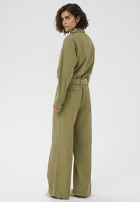 Femme aux cheveux courts et bouclés portant une combinaison longue olive à manches longues avec des jambes larges, se retournant sur l'épaule contre un fond uni.