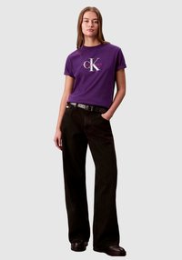 Fioletowy bawełniany T-shirt z białym i czerwonym logo, zestawiony z czarnymi, szerokimi spodniami z wysokim stanem oraz czarnymi butami. Prostota i casualowy styl.