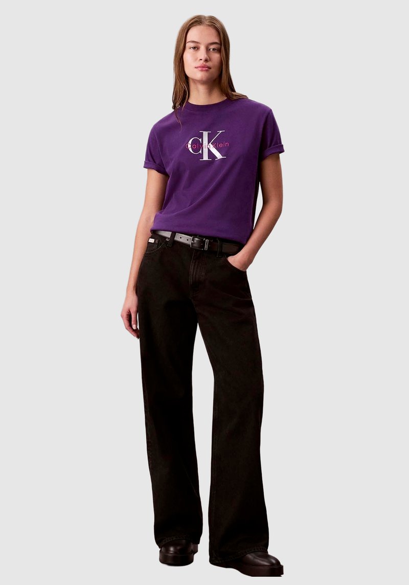 Fioletowy bawełniany T-shirt z białym i czerwonym logo, zestawiony z czarnymi, szerokimi spodniami z wysokim stanem oraz czarnymi butami. Prostota i casualowy styl.