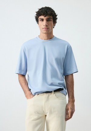 Giovane uomo con capelli corti e ricci, indossa una camicia a maniche corte azzurro chiaro e pantaloni beige, in piedi contro uno sfondo bianco uniforme.