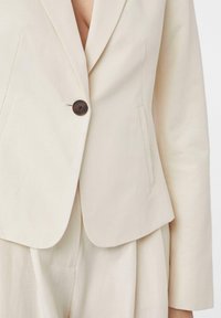 Beżowa marynarka z fakturowanej tkaniny, z pojedynczym ciemnym guzikiem i dopasowanym krojem. Posiada klapy oraz boczne kieszenie dla większej funkcjonalności.