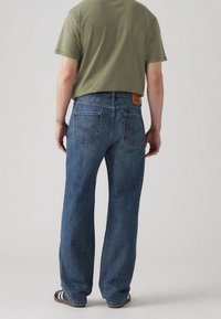 Blå denim jeans med rak benklipp, synliga sömmar och bakfickor. Bärs tillsammans med en olivgrön t-shirt och svarta sandaler.