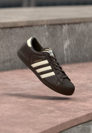 Czarny sneaker Adidas Superstar z białymi paskami i wzmocnionym noskiem, unoszący się nad betonową powierzchnią blisko kamiennego krawędzi.