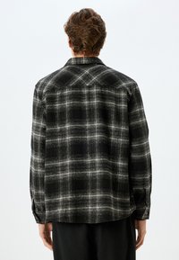 Camicia in flanella a quadri bianco e nero con vestibilità rilassata, maniche lunghe, polsini con bottoni e colletto standard; tessuto testurizzato con un motivo prominente.