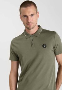 Olijfgroene polo met korte mouwen, gemaakt van zachte stof, met een drieknoopsluiting, contrasterende witte kraagaccenten en een rond logo.