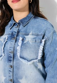 Chaqueta de mezclilla con tela azul clara, patrones de estrellas, bordes de los bolsillos deshilachados, embellishments de perlas y cierre de botón clásico.