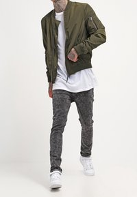 Olive green bomber jacket with a zip closure and side pockets, worn over a white T-shirt. Pair of grey textured skinny jeans and white sneakers.