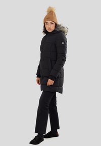 Zwarte pufferjas met een met bont gevoerde capuchon, ritssluiting en twee zijzakken. Gecombineerd met donkere jeans en zwarte sneakers, en afgewerkt met een tan beanie.