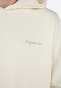 Cream-colored hoodie made of soft fabric, featuring a front logo "Pegador Co." in light beige, with a relaxed fit and a hood.