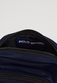 Interno di uno zaino in tessuto blu navy con foderatura nera. Presenta un'etichetta visibile con la scritta "POLO SPORT RALPH LAUREN." Vengono mostrati scomparti con zip.