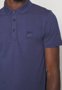 Homme portant un polo à manches courtes bleu marine avec trois boutons et un logo "BOSS" brodé sur le côté gauche de la poitrine.