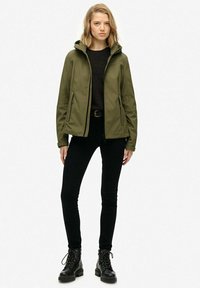 Młoda kobieta stoi, ubrana w oliwkową, kapturową kurtkę, czarny sweter, czarne spodnie i czarne botki na sznurówki, na jednolitym tle.