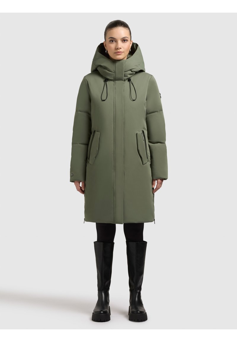 Olivgrüne gesteppte Parka mit Kapuze, Frontreißverschluss, Seitentaschen und Kordelzügen. Tragen über schwarzen Leggings und kniehohen schwarzen Stiefeln.